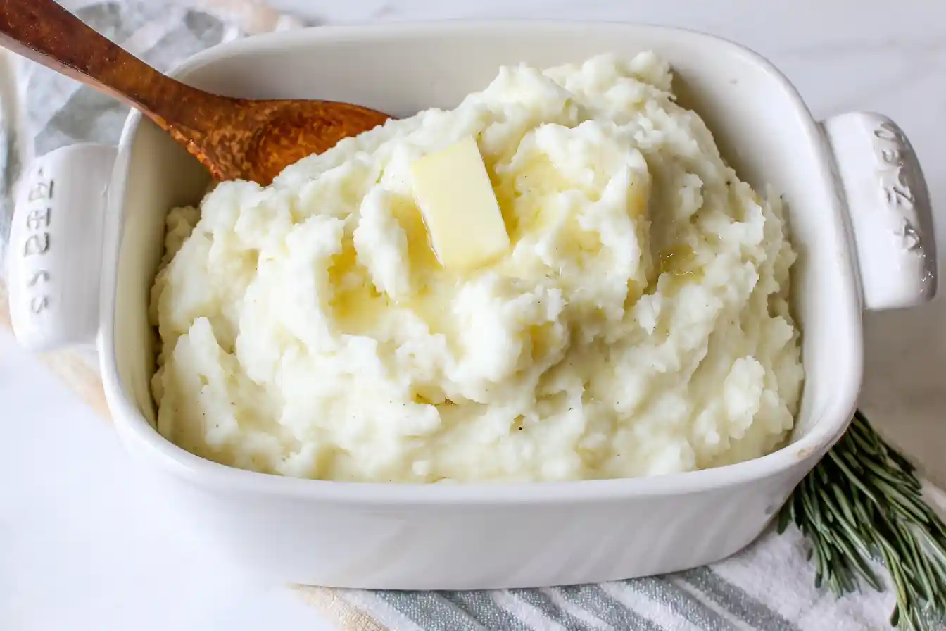 A close-up view of a bowl of fluffy Homemade Mashed Potatoes, topped with a dollop of melting butter and a sprinkle of fresh green chives.
