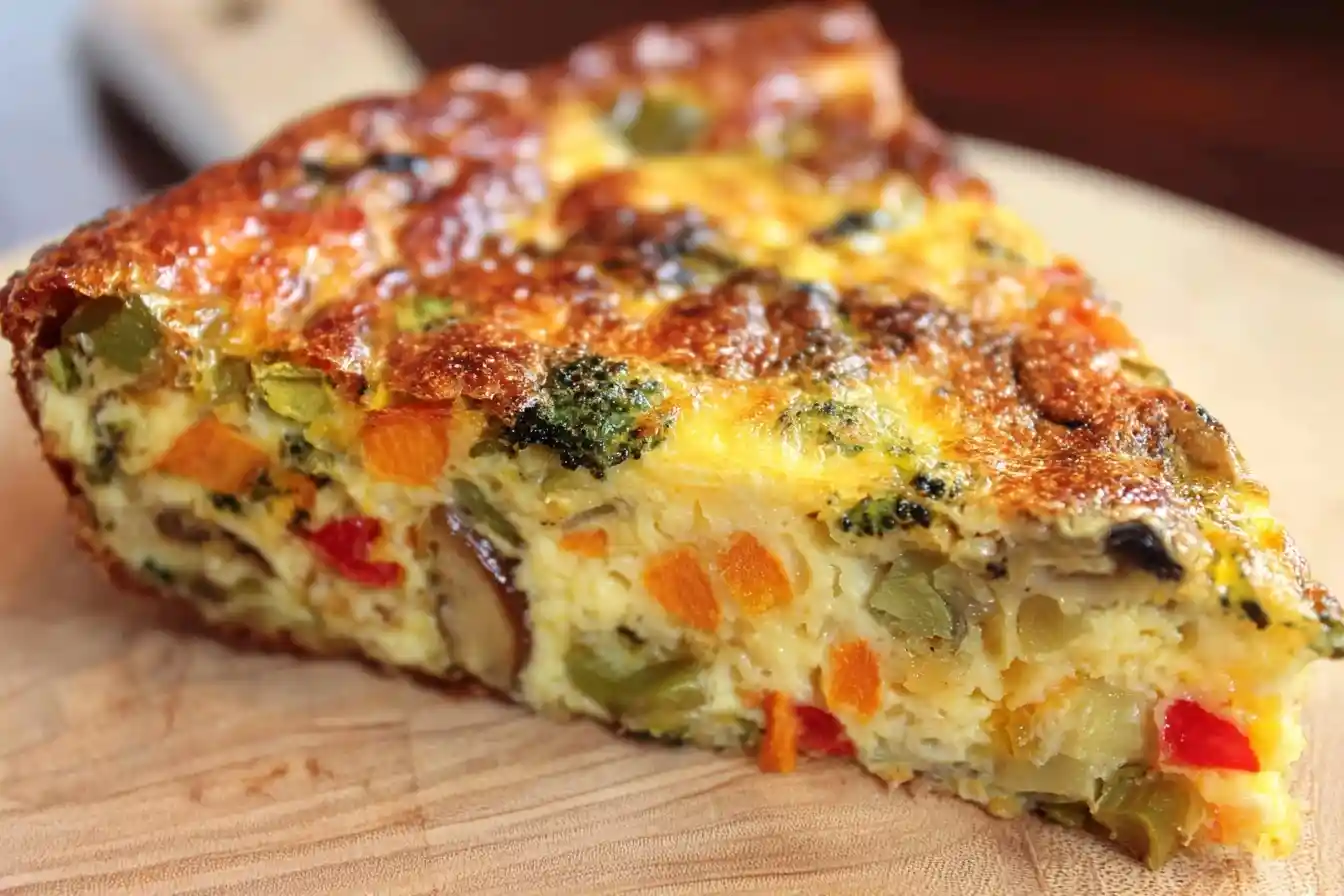 A close-up of a delicious slice of vegetable frittata packed with broccoli, carrots, and peppers on a wooden board.