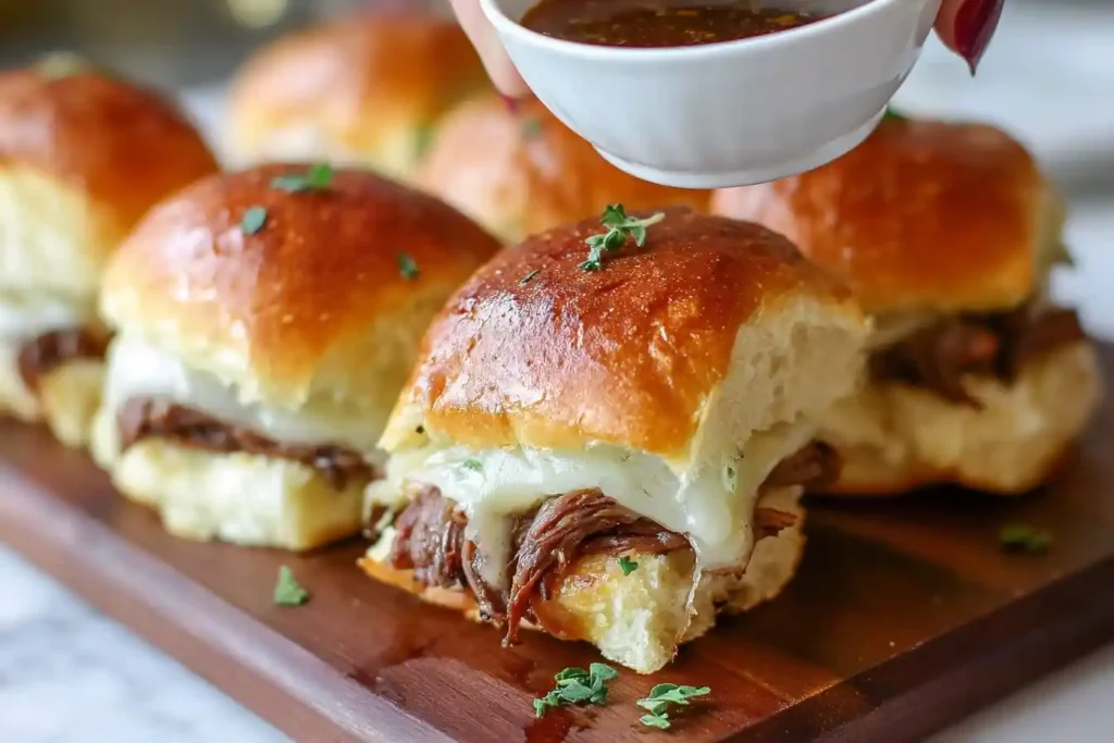 A platter of cheesy French Dip Sliders on a wooden board, with a small bowl of au jus for dipping.