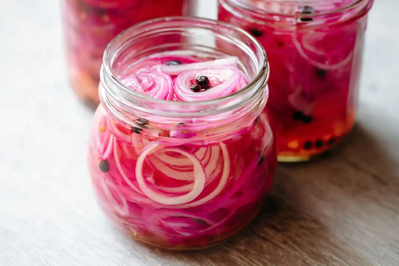 A close-up of a glass jar filled with vibrant, thinly sliced pickled red onions and black peppercorns on a wooden surface.