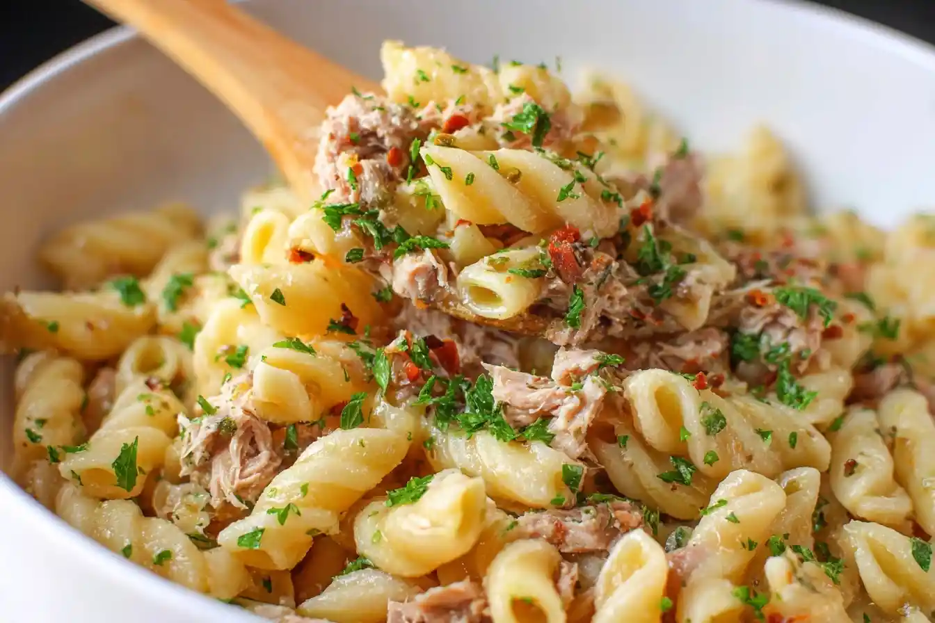 A close-up of a wooden spoon scooping up a serving of delicious tuna pasta salad from a white bowl.