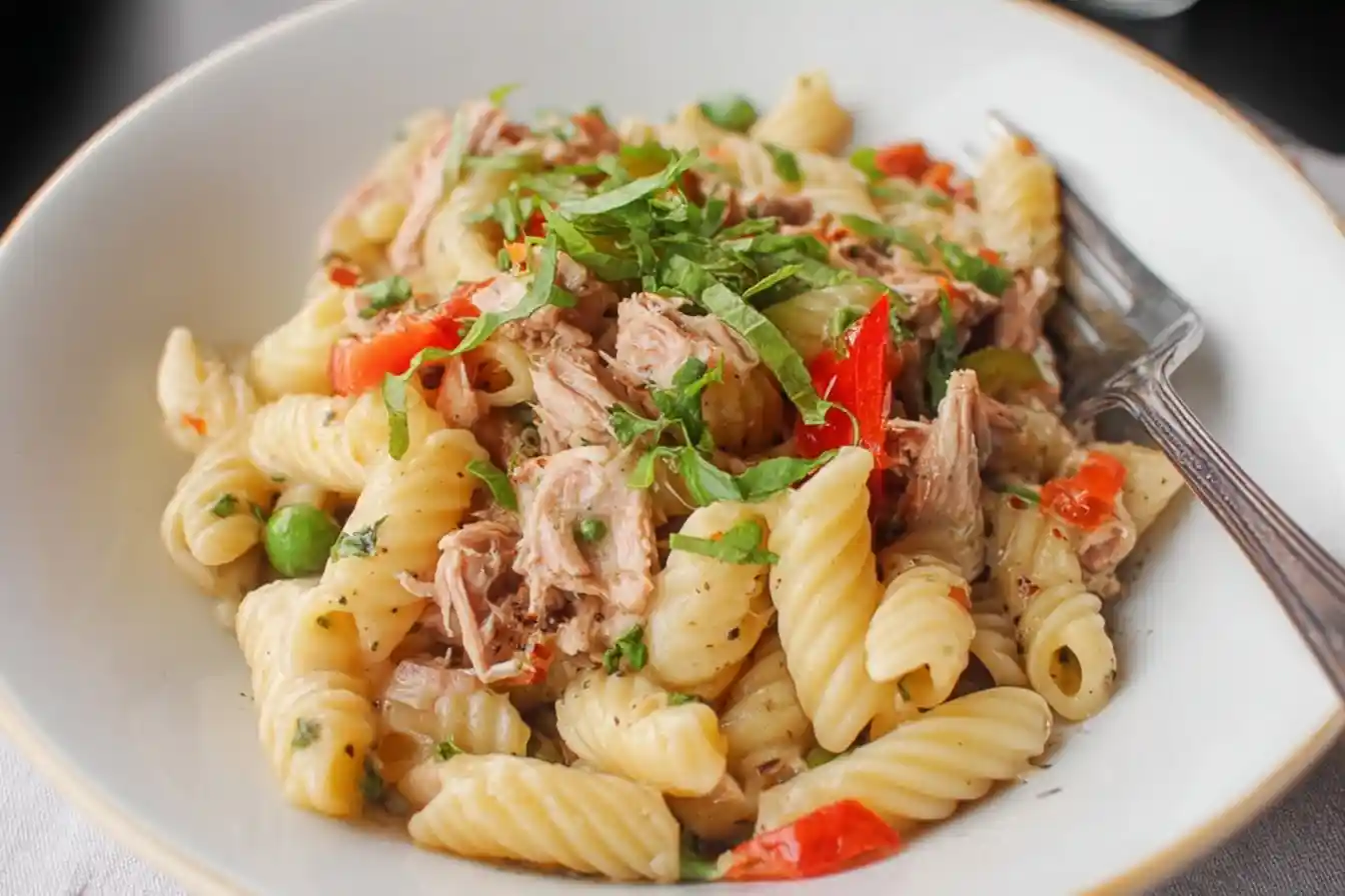 A close-up of a white bowl filled with a creamy tuna pasta salad, mixed with peas, red peppers, and topped with fresh herbs.