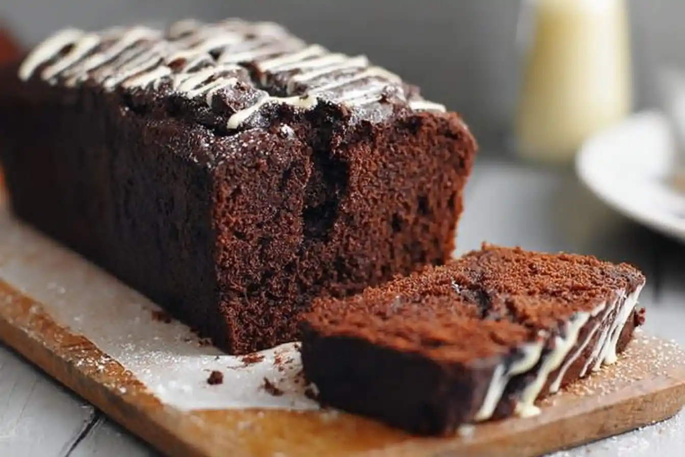 A rich chocolate loaf cake drizzled with white icing, with one slice cut and resting on a wooden board.
