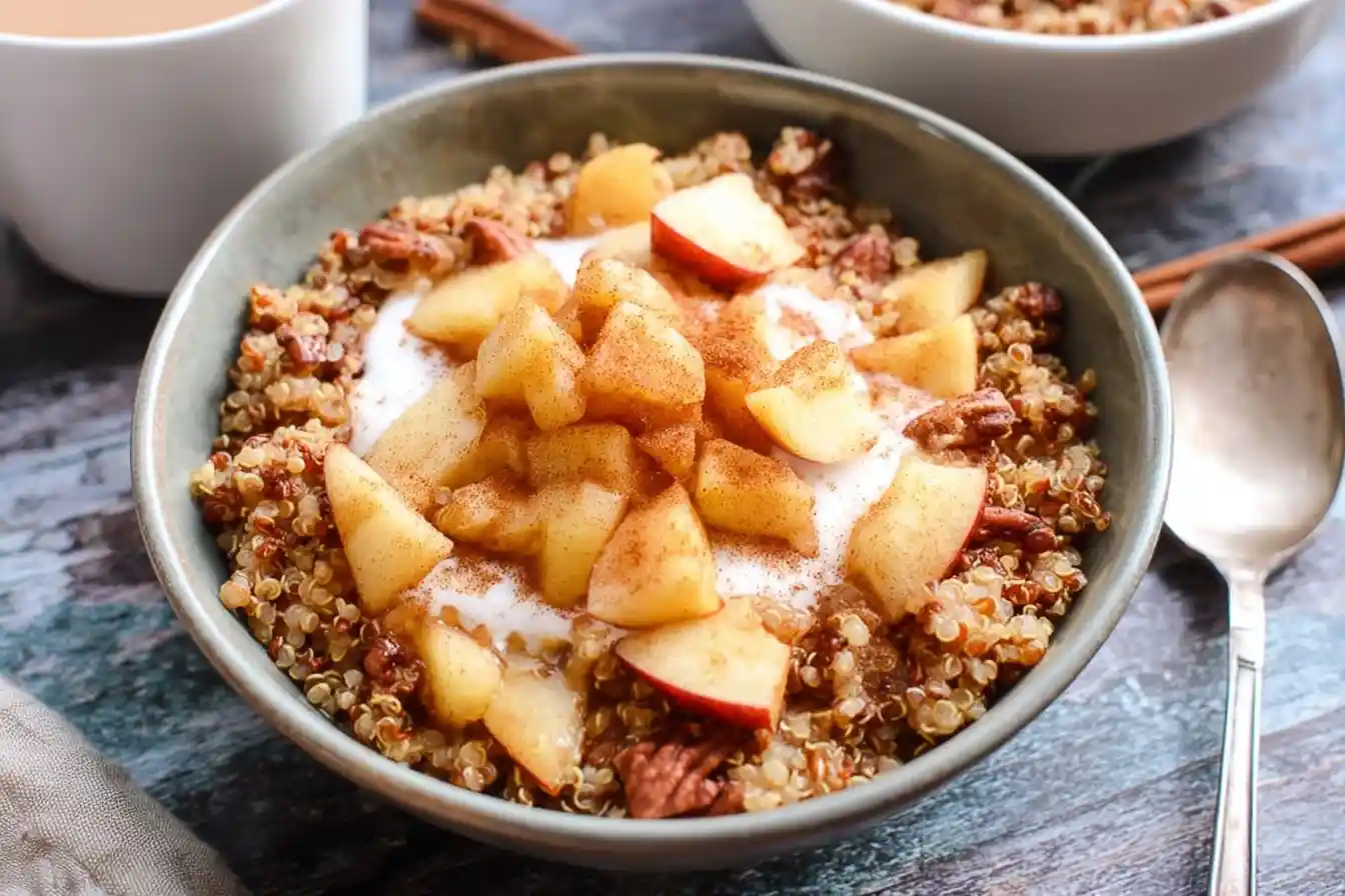 A close-up of a warm quinoa breakfast bowl topped with cinnamon apples, pecans, and a dollop of yogurt.