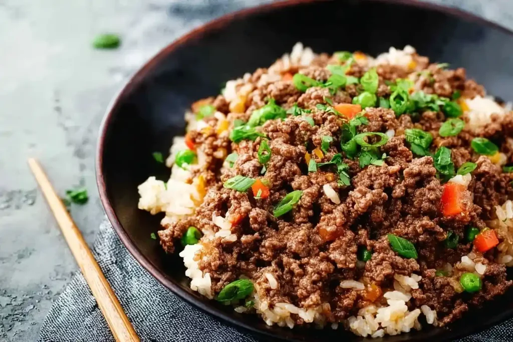 A close-up shot of a savory bowl of ground beef and rice, garnished with chopped green onions, peas, and diced red peppers.