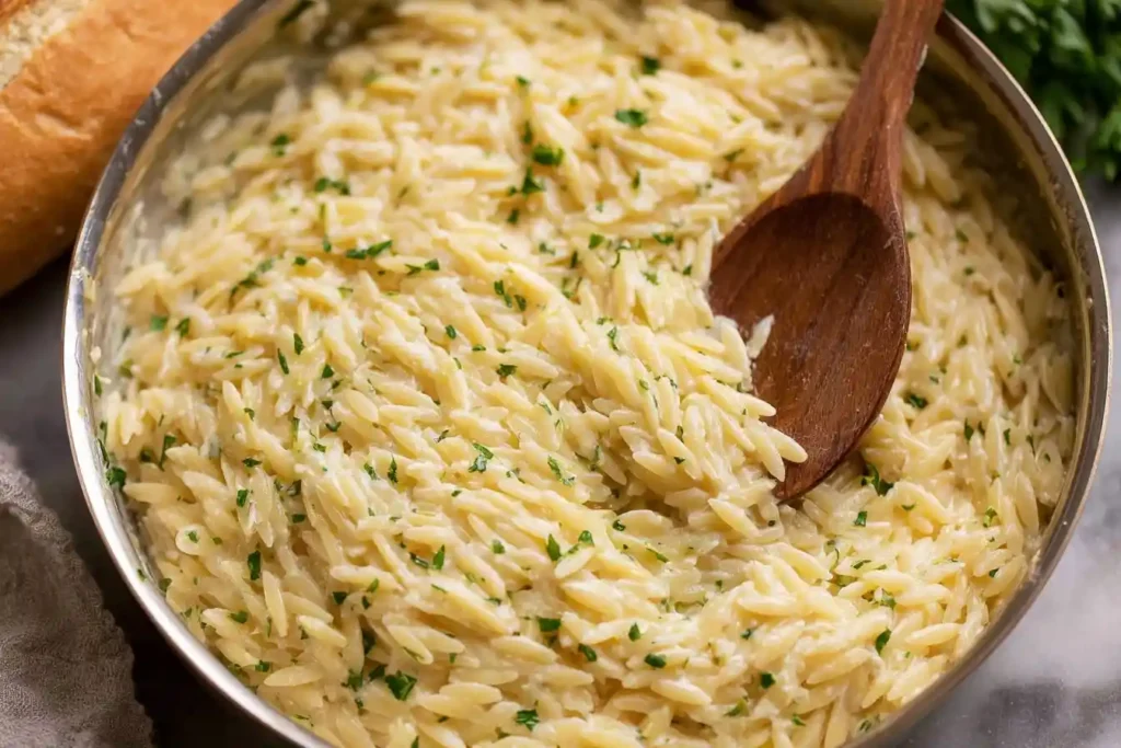 A close-up of a skillet filled with Creamy Orzo pasta, garnished with fresh parsley and being stirred with a wooden spoon.