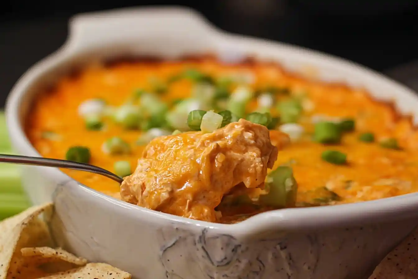 A close-up shot of a spoon scooping creamy Buffalo Chicken Dip out of a white baking dish, garnished with green onions.
