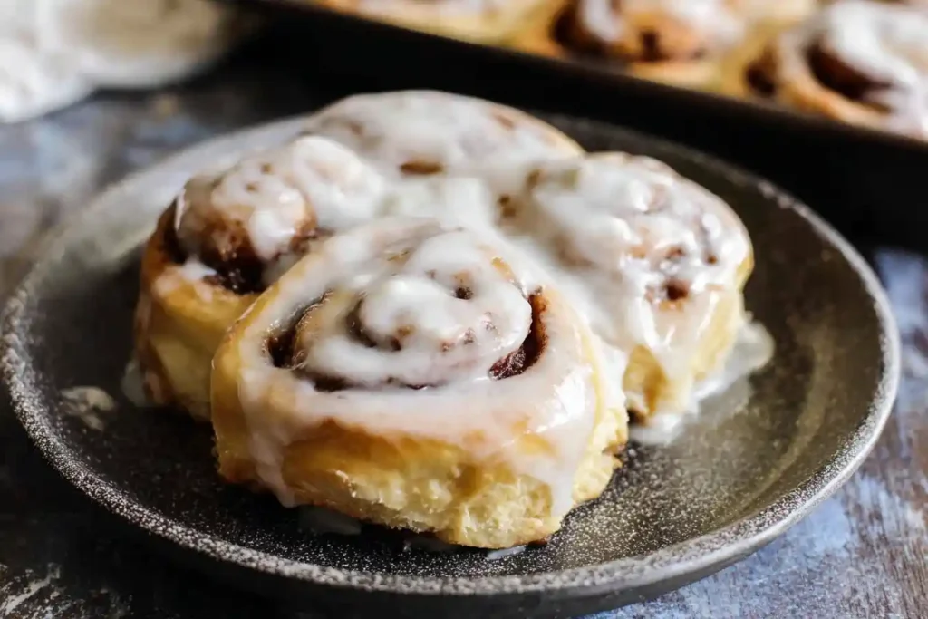 A close-up of several fluffy overnight cinnamon rolls on a dark plate, generously drizzled with white icing.