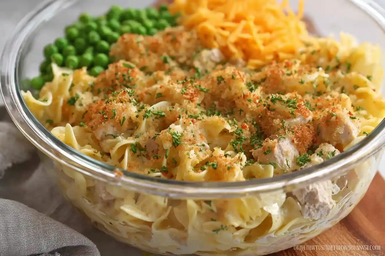 Glass bowl filled with creamy chicken noodle casserole, green peas, and shredded cheddar cheese.